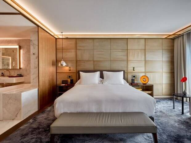 A modern Junior Suite features a double bed with white bedding, bedside tables, a padded headboard wall, and a view of the bathroom area separated by glass.